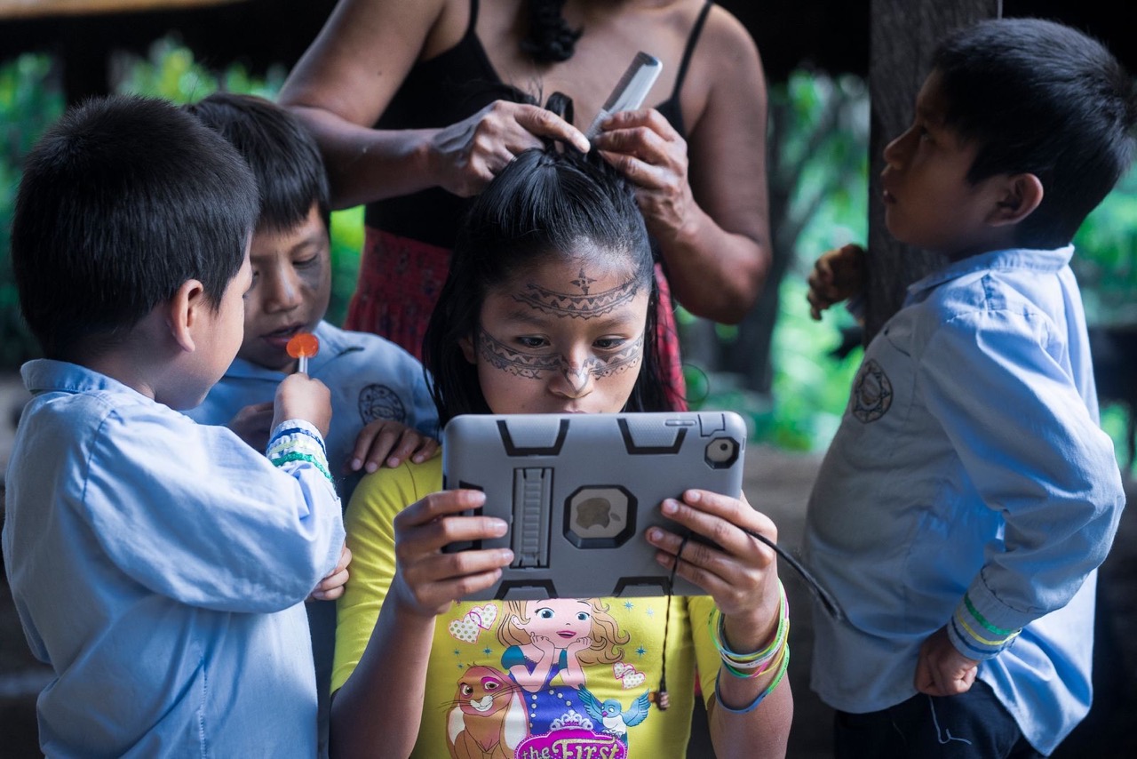 Leyli Gualinga joue sur un iPad pendant que sa mère Marcia Gualinga lui coiffe les cheveux et que son frère la regarde. - ©Misha Vallejo Leyli Gualinga joue sur un iPad pendant que sa mère Marcia Gualinga lui coiffe les cheveux et que son frère la regarde. - ©Misha Vallejo